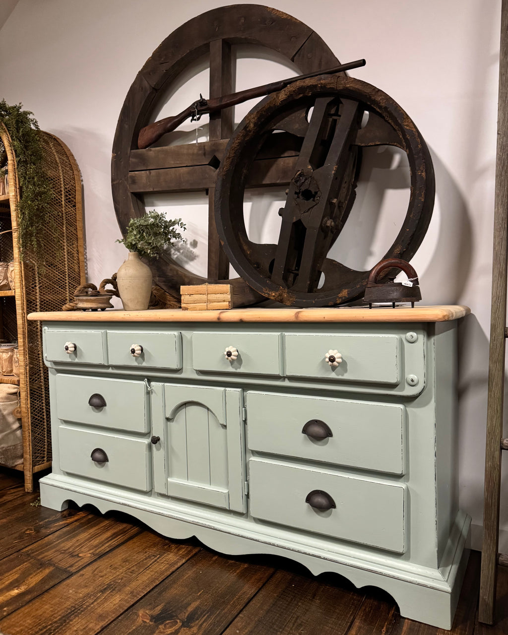 A distressed sage green wooden dresser with a light wood top, styled in a rustic room setting with vintage wheel decor and a rifle on top.