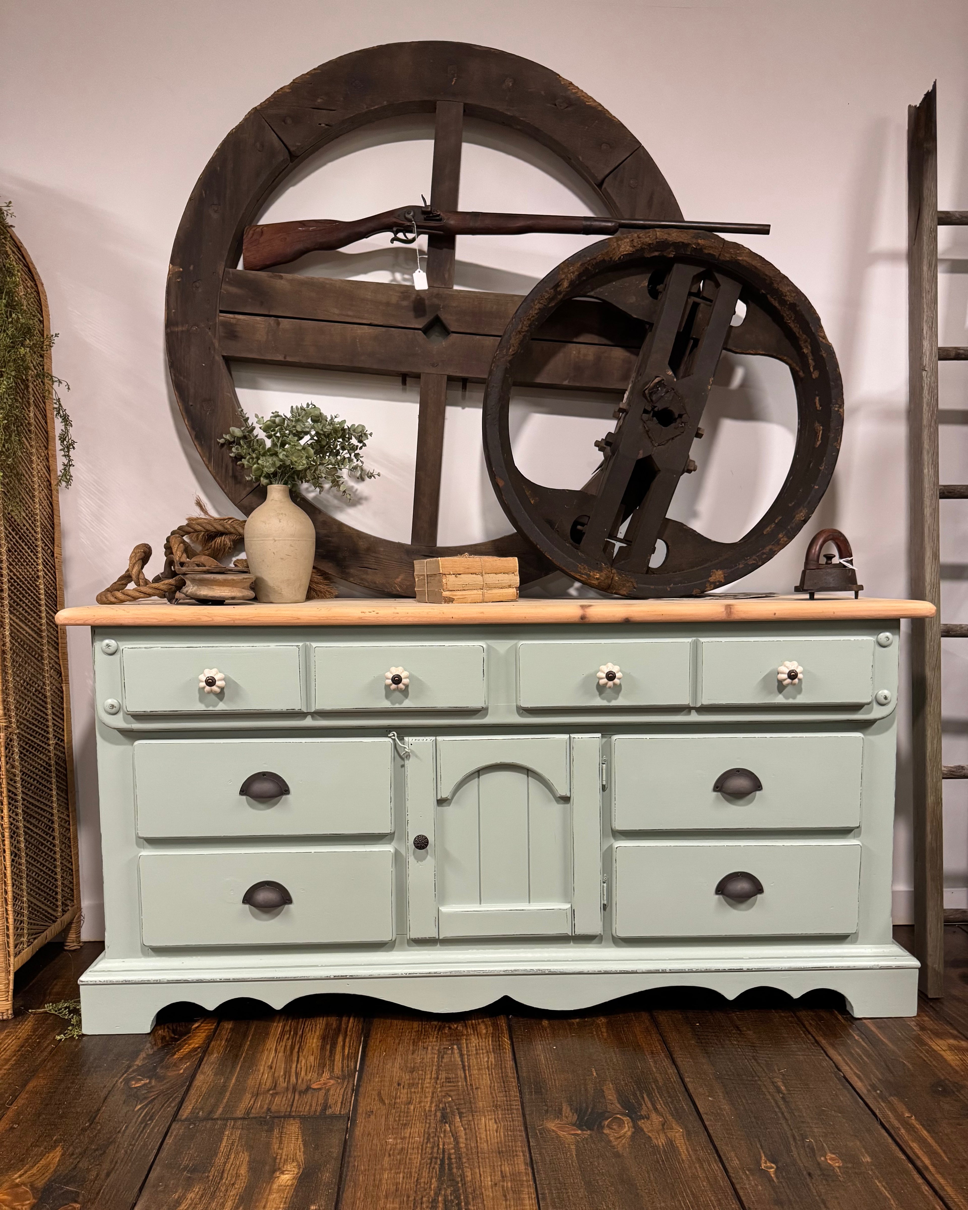 A vintage-style painted wooden dresser or sideboard with a light sage green body, a natural wood top, and dark metal drawer pulls, styled in a rustic room with large wooden wheels and a rifle behind it.