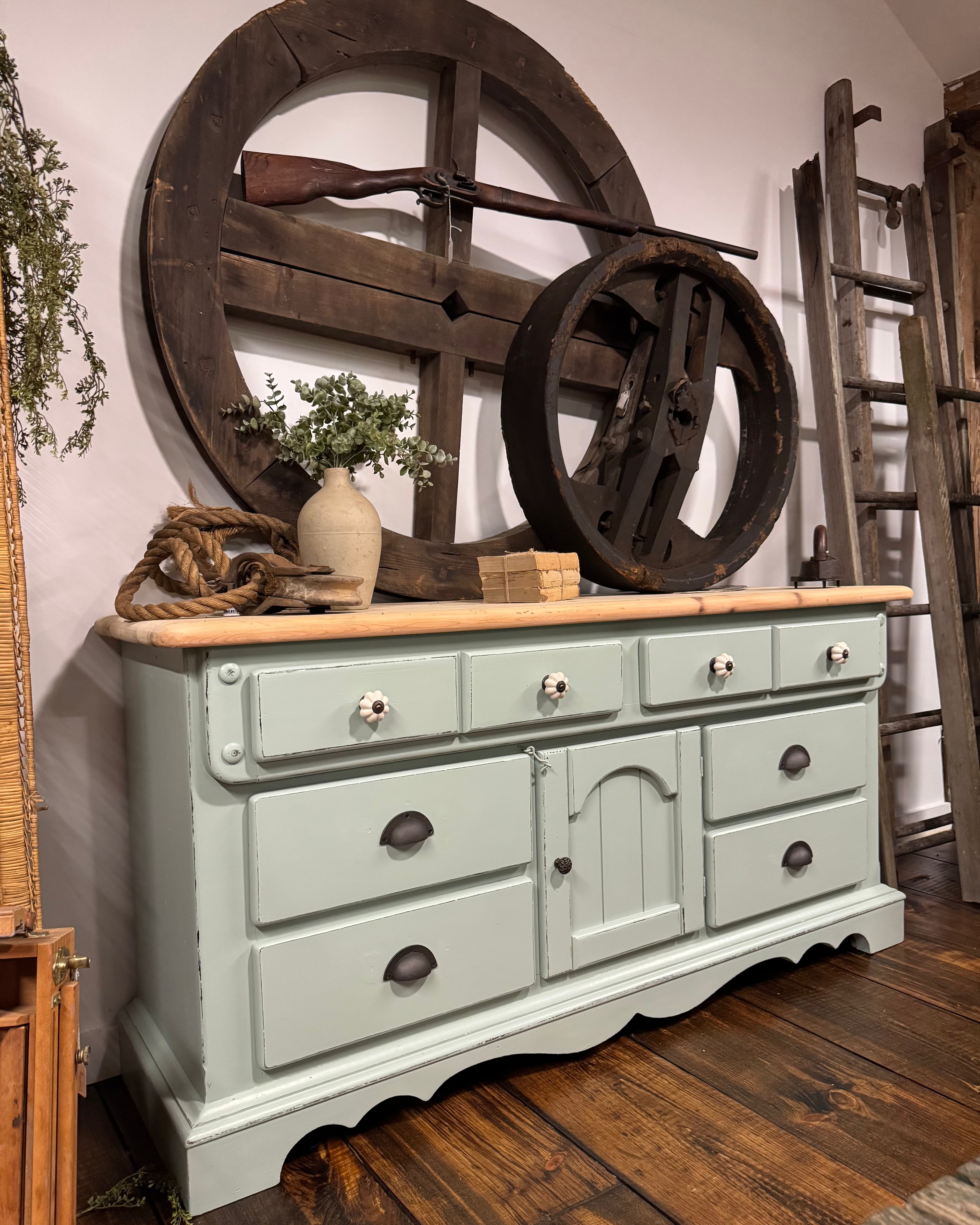 A distressed sage green wooden dresser with a light wood top, styled in a rustic room setting with vintage wheel decor and wooden ladders in the background.