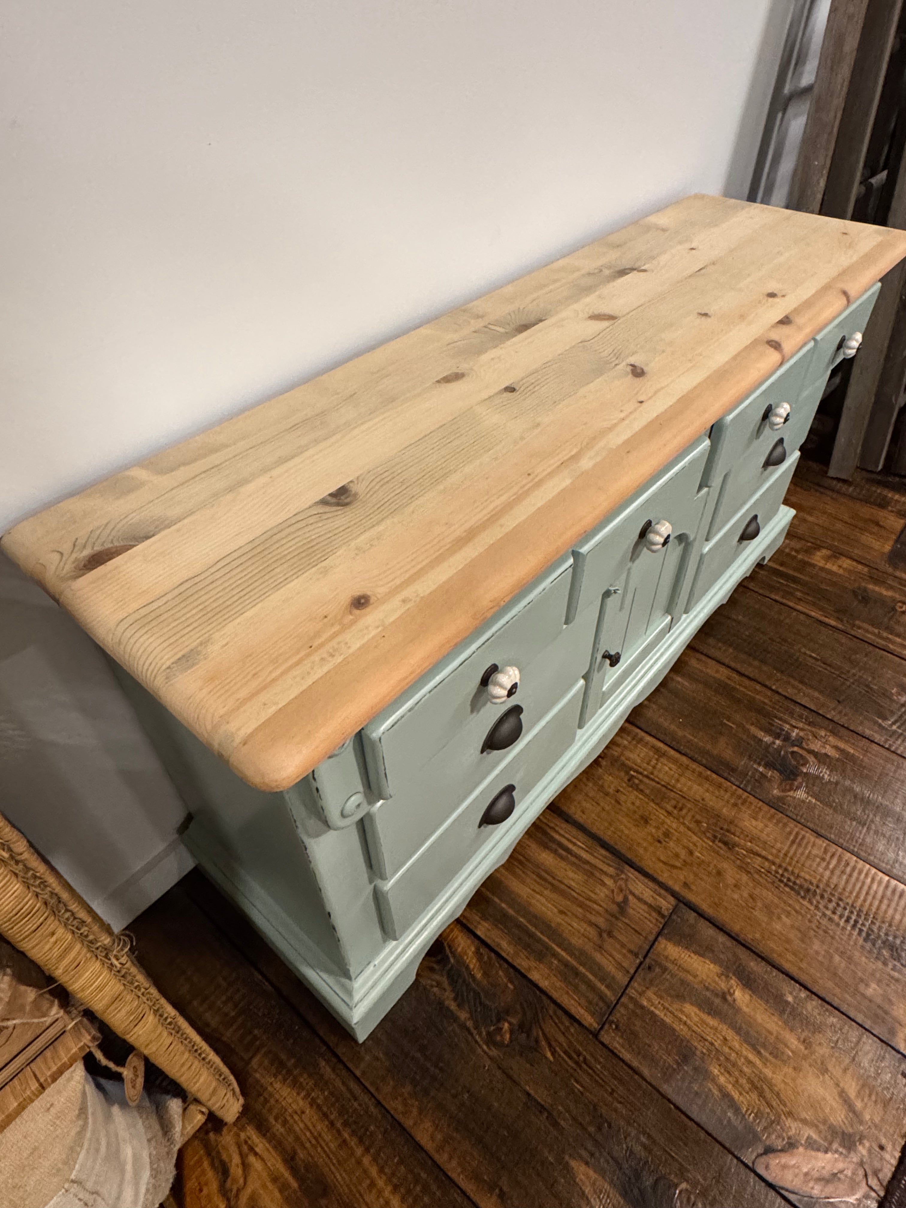 A rustic pine wood chest with a sage green painted frame and drawer fronts, photographed in a home setting on a dark wood floor against a plain wall.
