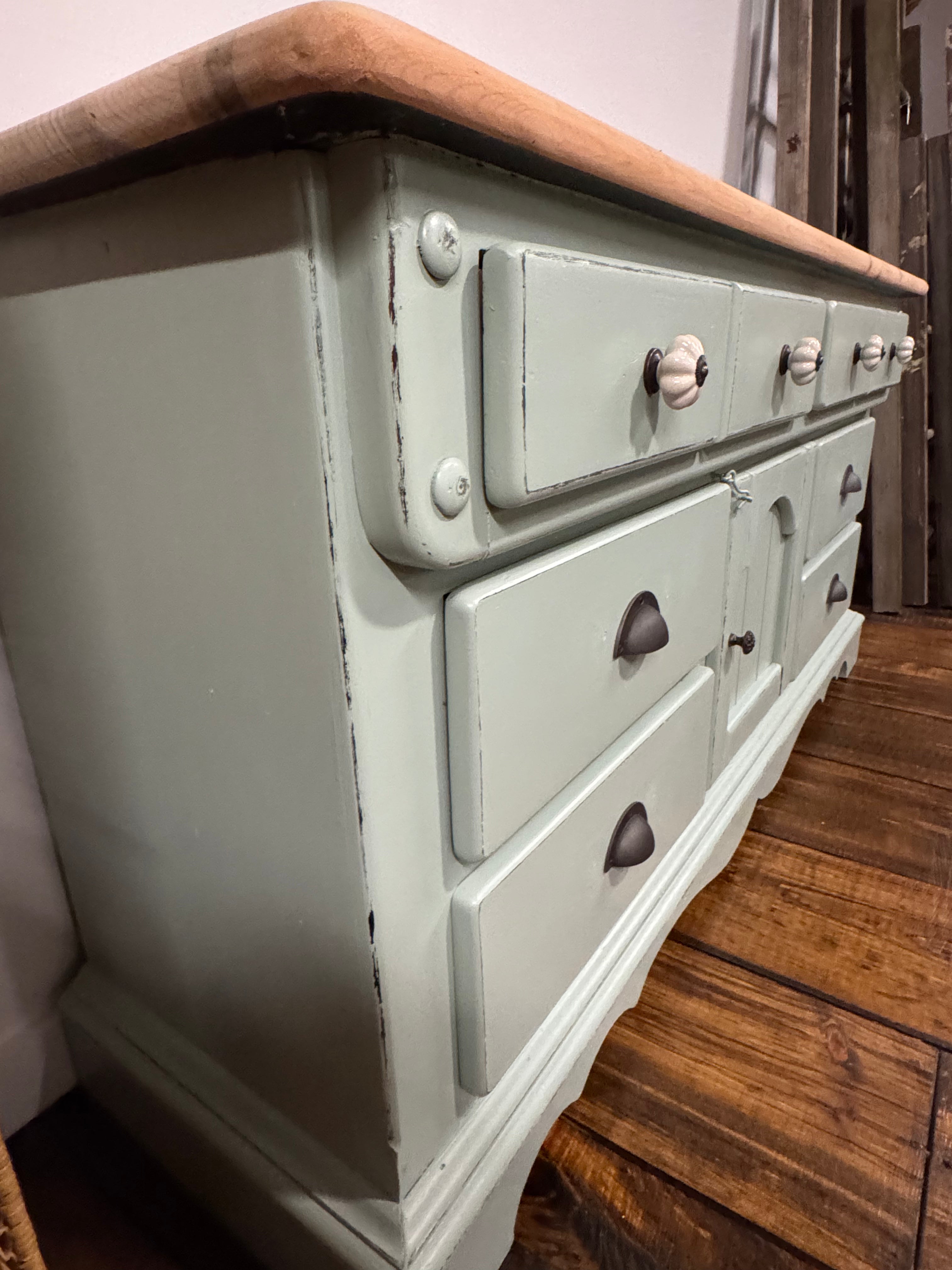 A distressed sage green wooden dresser with a natural wood top and white knobs, shown in a home setting on a wooden floor.