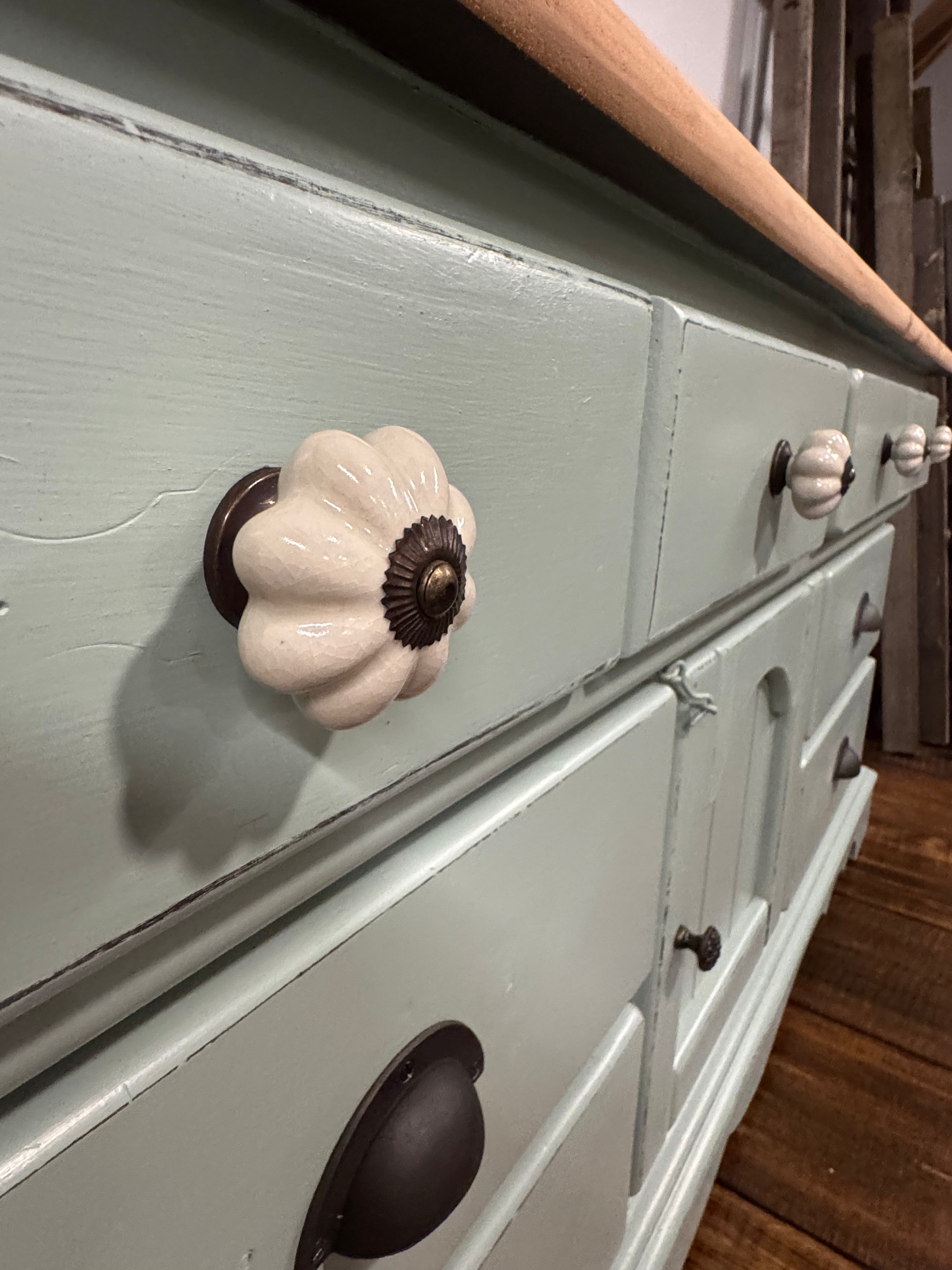 A close-up of a painted wooden dresser with a sage green finish, featuring a large cream-colored flower-shaped drawer knob and smaller dark metal knobs, set against a dark wood background.