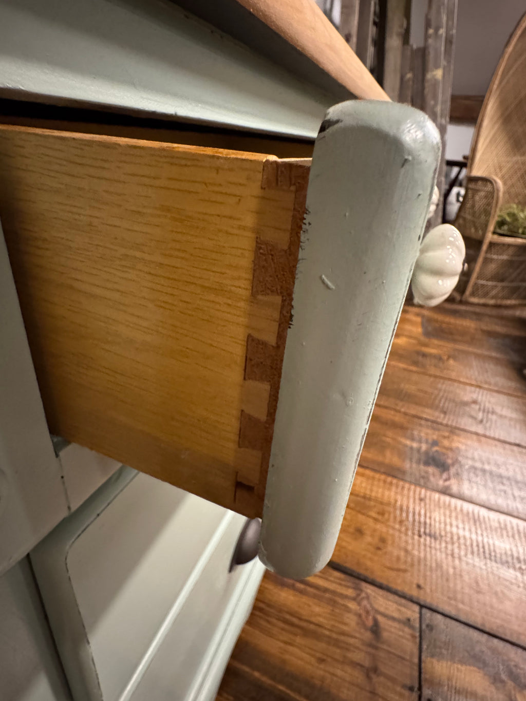 A close-up of a vintage-style wooden cabinet drawer with a light natural wood interior, a white-painted front, and a round white knob, set against a rustic wooden floor background.