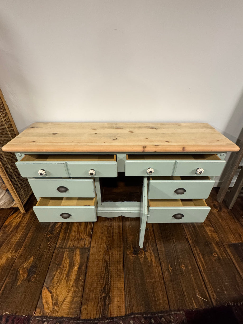 A rustic wooden console table with a sage green painted frame and six drawers, shown with several drawers open on a dark wood floor against a plain wall.
