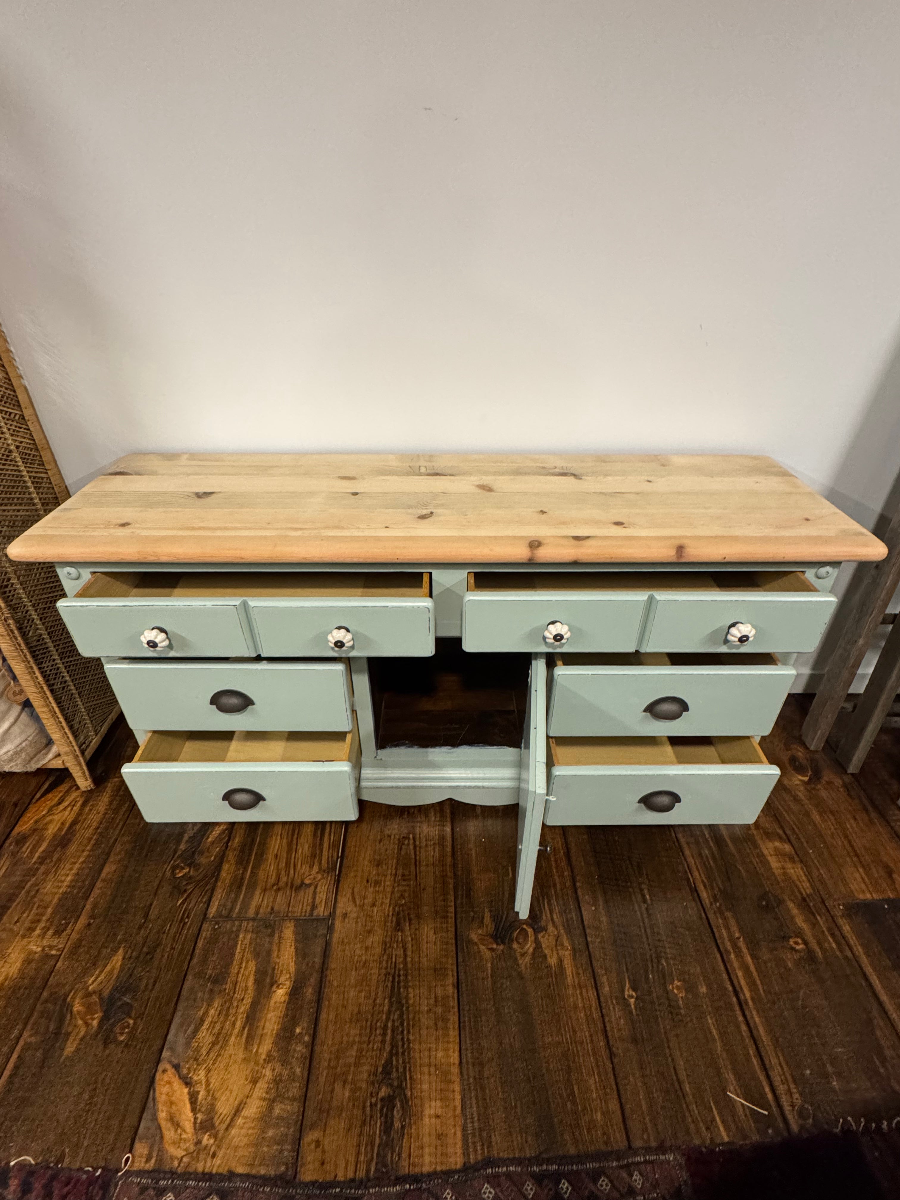 A rustic wooden console table with a sage green painted frame and six drawers, shown with several drawers open on a dark wood floor against a plain wall.